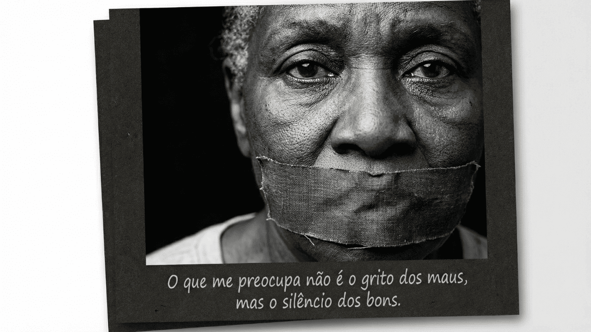 Retrato dramático e artístico em preto e branco de um rosto com a boca tapada por uma fita, simbolizando a censura e a omissão. Abaixo, a poderosa frase de Martin Luther King sobre cumplicidade: 'O que me preocupa não é o grito dos maus, mas o silêncio dos bons'.