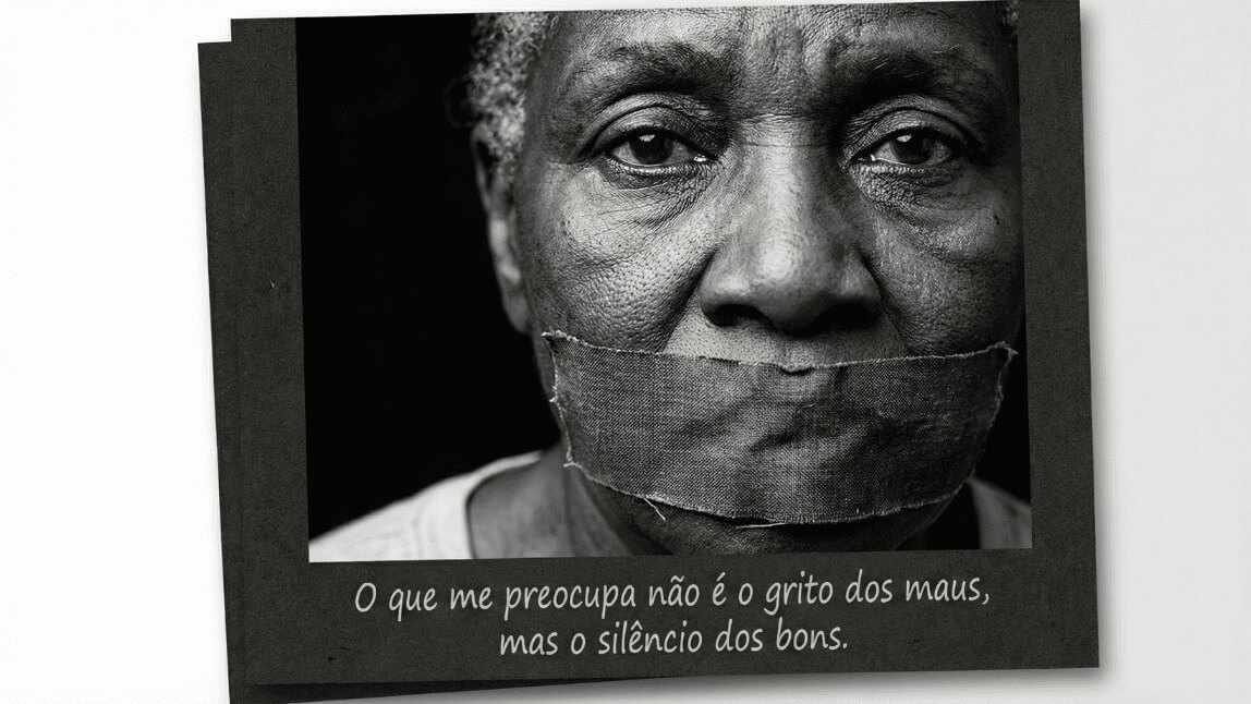 Retrato preto e branco de um rosto com a boca tapada por uma fita, simbolizando a censura e a omissão. Abaixo frase: 'O que me preocupa não é o grito dos maus, mas o silêncio dos bons'.