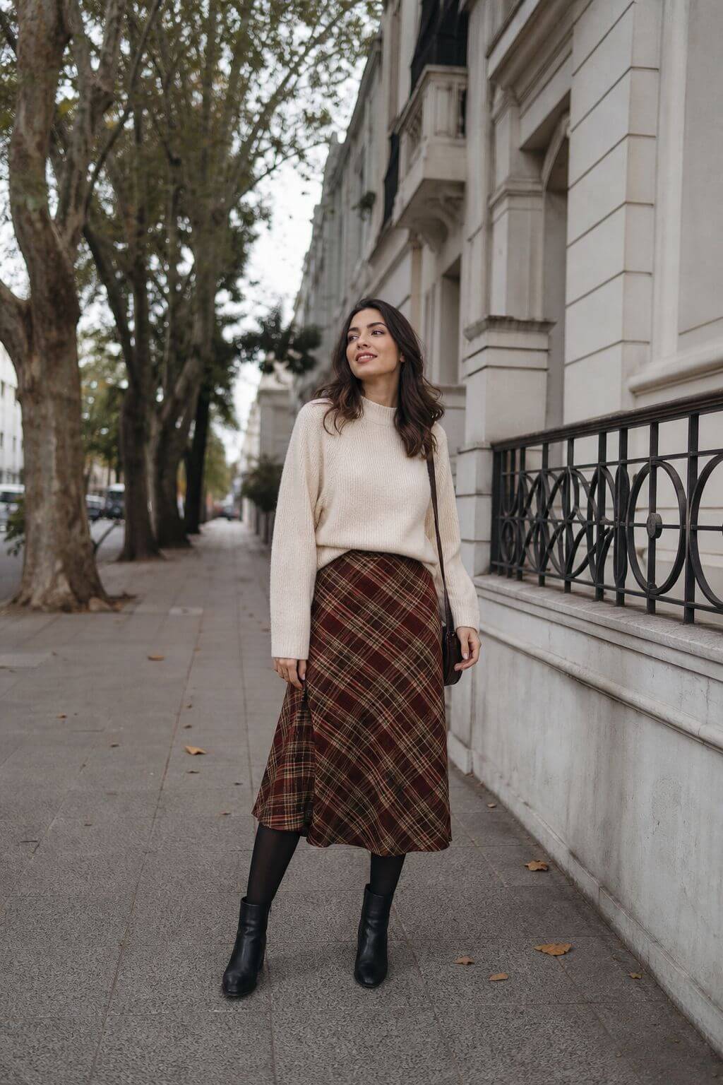 Look feminino de inverno com suéter creme, saia midi xadrez, meia-calça preta e botas de cano curto.