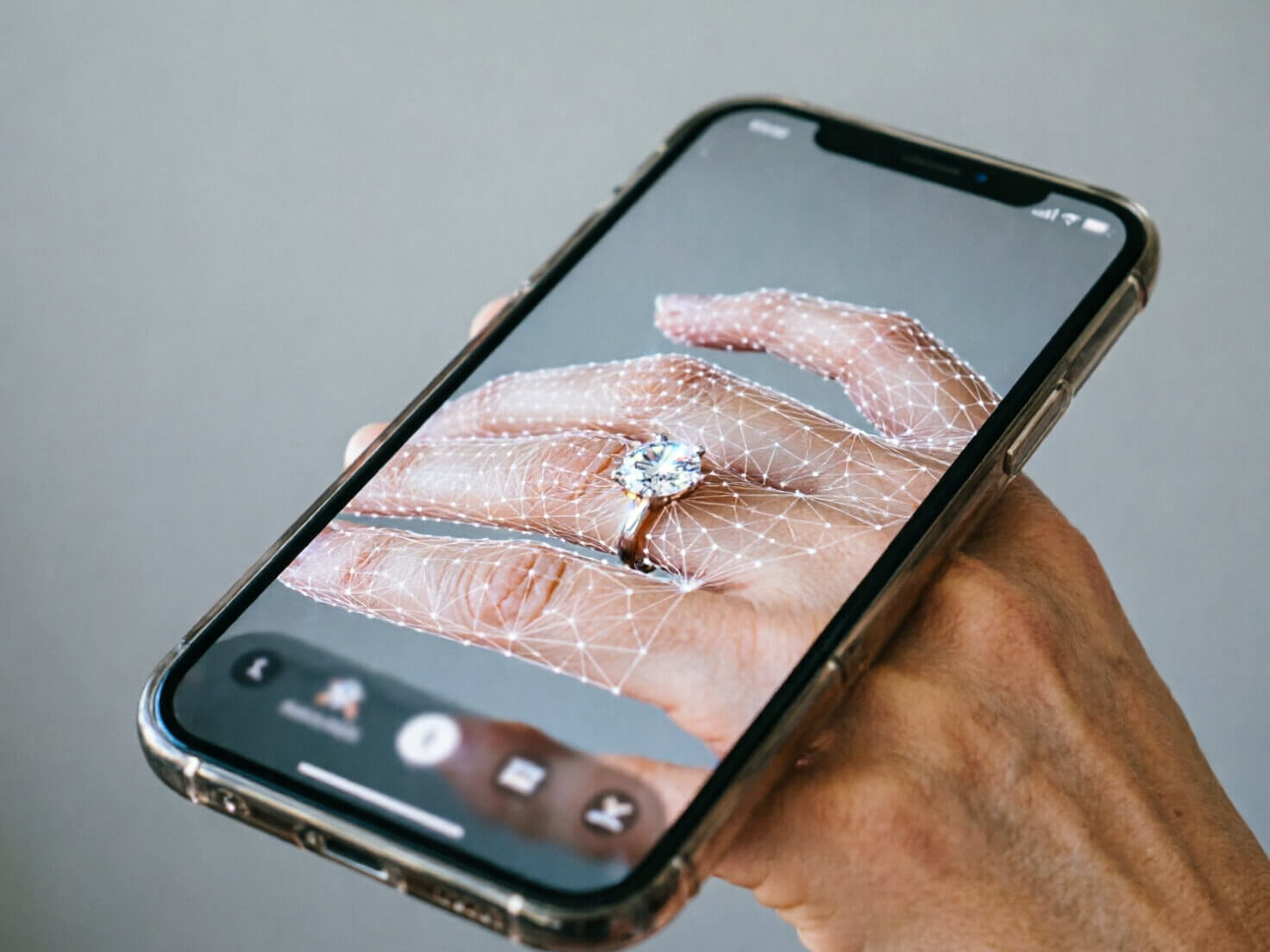 What Is a Virtual Ring Try-On? Photorealistic close-up of a smartphone screen held over a bare hand, showing an AR jewelry app interface mid-scan with a subtle white hand-tracking mesh/dot overlay on the fingers and a sparkling solitaire diamond ring rendered onto the ring finger, app UI elements (thumbnail ring carousel at bottom) visible but blurred for realism, bright clean studio lighting