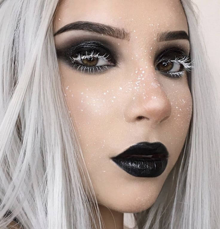 Maquiagem gótica com glitter prateado, cílios claros, lábios pretos e pele matte