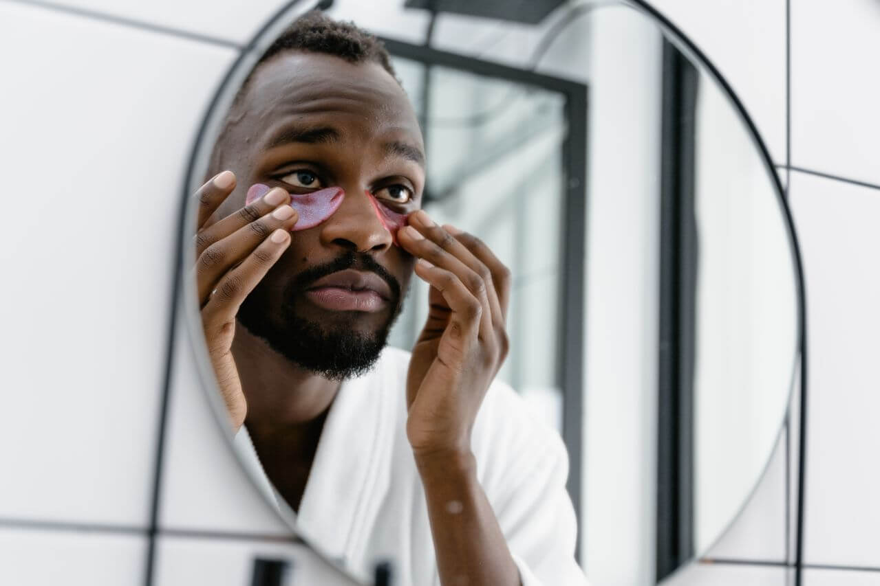 Man in white bathrobe applying eye patches in bathroom mirror for skincare routine. Man in white bathrobe applying eye patches in bathroom mirror for skincare routine.