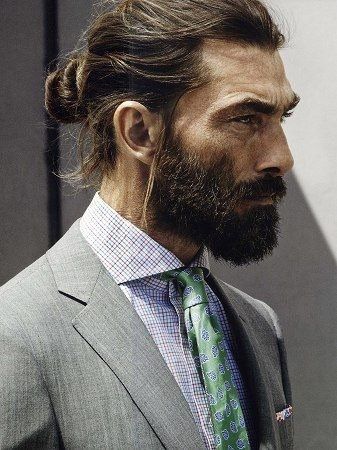 Mann mit zusammengebundenem Dutt und Vollbart, im Anzug getragen, moderner Low Bun als Langhaarfrisur für Männer.